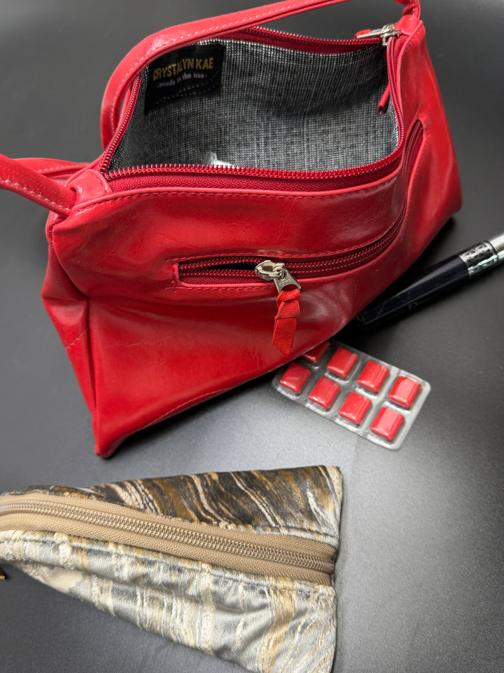 Red leather pouch with gold zipper on a gray surface, next to a camouflage pouch and red items.