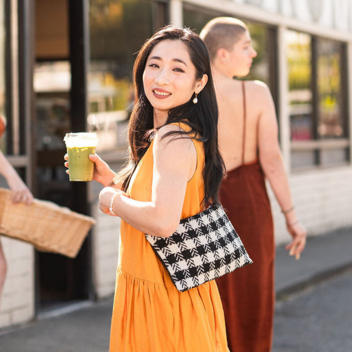 Woman in a yellow dress holding a green cup wearing a houndstooth bag outside a store with other people in the background.#color_gothic-houndstooth