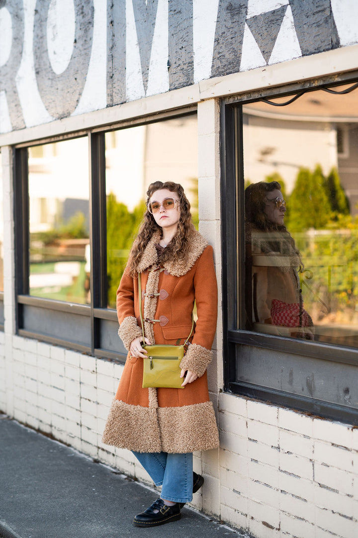 Woman in a brown coat standing in front of a building with geometric window design#color_citrine