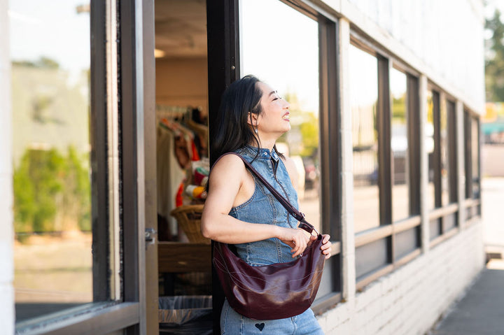 Woman holding a brown leather bag standing outside a store.#color_wine