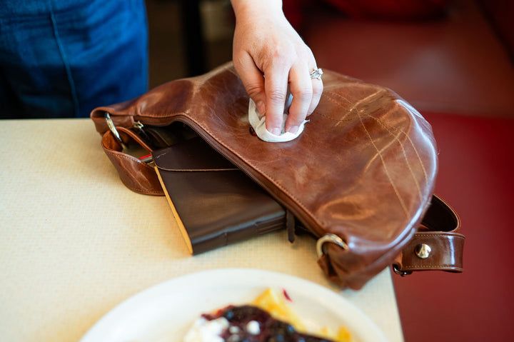 Person cleaning a brown leather bag with a white cloth on a table.#color_ale-brown