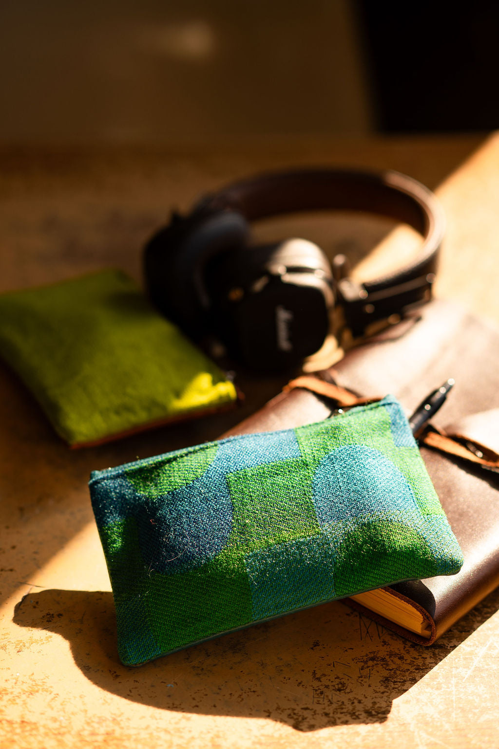 Green checkered pouch on a textured surface with headphones and a pen in the background#color_sky-arches-teal-green
