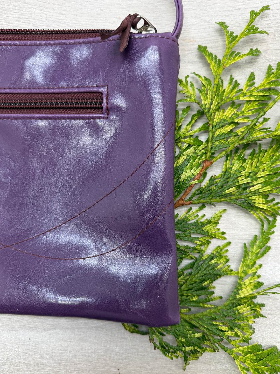 Purple leather crossbody bag with a textured surface on a light wooden background with green leaves.#color_plum