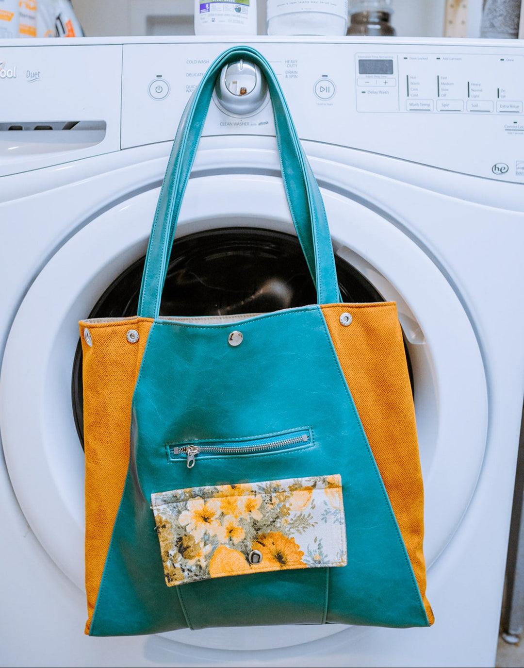 machine-wasahble Turquoise and orange tote bag with floral pocket hanging on a washing machine.